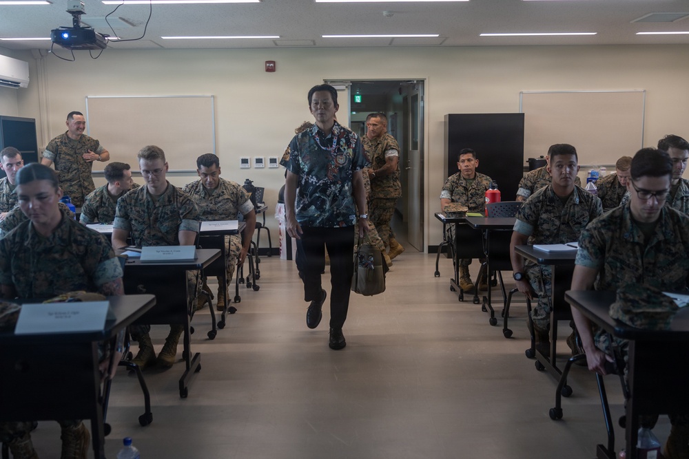 A civilian instructor or guest speaker in a Hawaiian/floral shirt walks down the center aisle of a military classroom filled with uniformed personnel in camouflage uniforms. The setting appears to be a formal training or briefing environment, likely on a U.S. military installation. Students are seated at individual desks with laptops and materials, suggesting an active educational session.