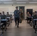 A civilian instructor or guest speaker in a Hawaiian/floral shirt walks down the center aisle of a military classroom filled with uniformed personnel in camouflage uniforms. The setting appears to be a formal training or briefing environment, likely on a U.S. military installation. Students are seated at individual desks with laptops and materials, suggesting an active educational session.