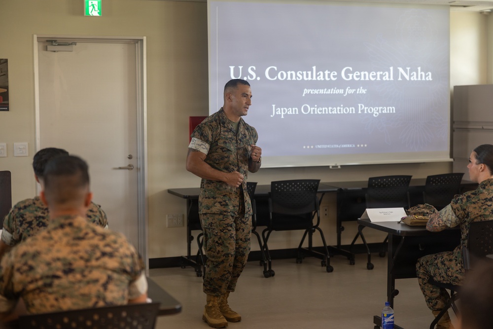 A U.S. Marine in camouflage uniform is presenting at a Japan Orientation Program briefing hosted by the U.S. Consulate General Naha. Several other Marines in uniform are seated as an audience in a classroom-style setting. A projected slide behind the speaker displays the title 'U.S. Consulate General Naha presentation for the Japan Orientation Program.'