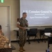 A U.S. Marine in camouflage uniform is presenting at a Japan Orientation Program briefing hosted by the U.S. Consulate General Naha. Several other Marines in uniform are seated as an audience in a classroom-style setting. A projected slide behind the speaker displays the title 'U.S. Consulate General Naha presentation for the Japan Orientation Program.'
