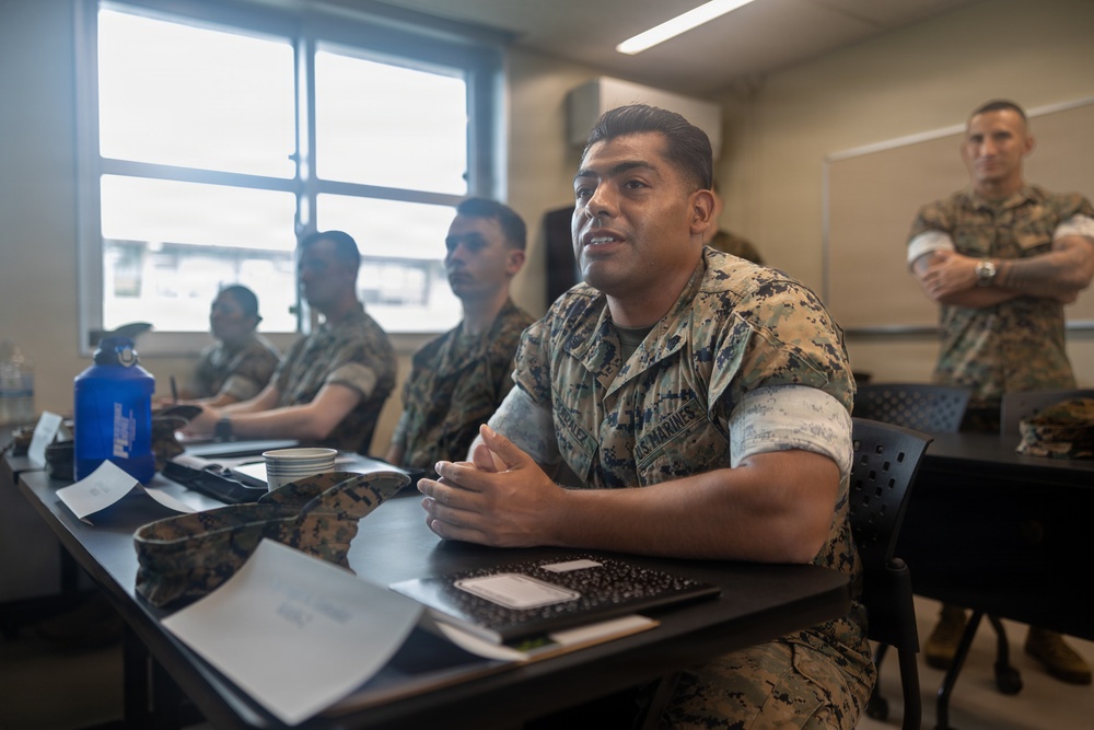A group of U.S. Marines in MARPAT camouflage uniforms are seated at desks in a classroom setting, appearing to be engaged in a briefing or training session. The foreground Marine appears to be speaking or responding to a question, with clasped hands resting on the desk. A senior Marine stands in the background with arms crossed, observing the session.