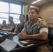 A group of U.S. Marines in MARPAT camouflage uniforms are seated at desks in a classroom setting, appearing to be engaged in a briefing or training session. The foreground Marine appears to be speaking or responding to a question, with clasped hands resting on the desk. A senior Marine stands in the background with arms crossed, observing the session.