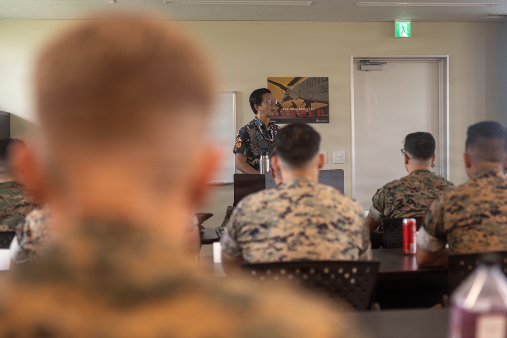 A civilian presenter or instructor is addressing a group of U.S. Marines in what appears to be a briefing or classroom setting. The Marines are seated at tables in military camouflage uniforms (MARPAT), listening to the speaker. A U.S. Marines recruitment or motivational poster is visible on the wall behind the presenter.