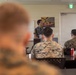A civilian presenter or instructor is addressing a group of U.S. Marines in what appears to be a briefing or classroom setting. The Marines are seated at tables in military camouflage uniforms (MARPAT), listening to the speaker. A U.S. Marines recruitment or motivational poster is visible on the wall behind the presenter.