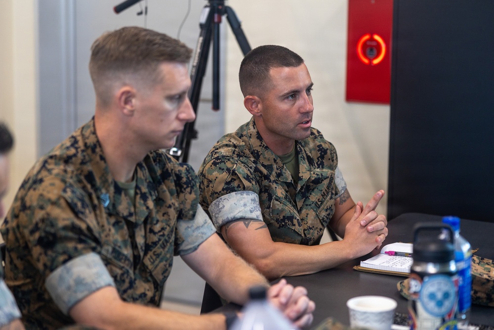 Two U.S. Marines in MARPAT camouflage uniforms are seated at a table engaged in what appears to be a briefing or discussion. The Marine on the right is actively speaking with clasped hands, while the one on the left listens attentively. The setting appears to be an indoor military facility or conference room.