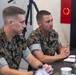 Two U.S. Marines in MARPAT camouflage uniforms are seated at a table engaged in what appears to be a briefing or discussion. The Marine on the right is actively speaking with clasped hands, while the one on the left listens attentively. The setting appears to be an indoor military facility or conference room.
