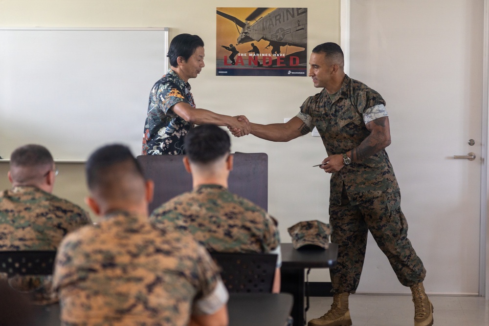 A civilian in a floral shirt is shaking hands with a U.S. Marine in woodland MARPAT camouflage uniform in what appears to be a briefing or classroom setting. Several other Marines in uniform are seated in the foreground with their backs to the camera. A U.S. Marines recruitment poster reading 'The Marines Have Landed' is visible on the wall behind them.