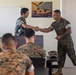 A civilian in a floral shirt is shaking hands with a U.S. Marine in woodland MARPAT camouflage uniform in what appears to be a briefing or classroom setting. Several other Marines in uniform are seated in the foreground with their backs to the camera. A U.S. Marines recruitment poster reading 'The Marines Have Landed' is visible on the wall behind them.