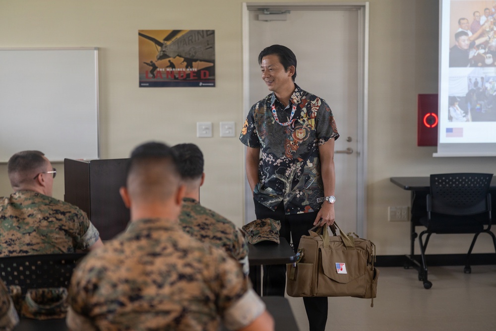 A civilian presenter in a decorative patterned shirt stands at the front of a classroom briefing U.S. Marines in camouflage uniforms. He is smiling and holding a tan tactical-style bag with what appears to be a small American flag patch. A projection screen displaying photos is visible in the background alongside a Marine Corps recruitment poster.