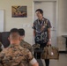 A civilian presenter in a decorative patterned shirt stands at the front of a classroom briefing U.S. Marines in camouflage uniforms. He is smiling and holding a tan tactical-style bag with what appears to be a small American flag patch. A projection screen displaying photos is visible in the background alongside a Marine Corps recruitment poster.