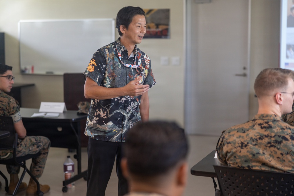 A civilian presenter wearing a decorative Japanese-style patterned shirt and lanyard is addressing a group of U.S. Marines in a classroom or briefing room setting. The Marines are seated at desks in military camouflage uniforms (MARPAT pattern), attentively listening to the speaker. The setting appears to be a formal briefing or cultural/educational presentation.