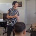 A civilian presenter wearing a decorative Japanese-style patterned shirt and lanyard is addressing a group of U.S. Marines in a classroom or briefing room setting. The Marines are seated at desks in military camouflage uniforms (MARPAT pattern), attentively listening to the speaker. The setting appears to be a formal briefing or cultural/educational presentation.