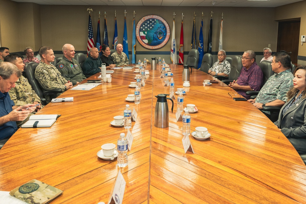 A formal meeting is taking place in a military conference room at Commander, Joint Region Marianas headquarters, as indicated by the official seal visible on the wall. Military personnel in various uniforms are seated on one side of a large wooden conference table, while civilian officials in business and aloha-style attire are seated on the opposite side. The setting suggests a high-level coordination or briefing between U.S. military leadership and local civilian government officials.
