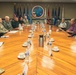 A formal meeting is taking place in a military conference room at Commander, Joint Region Marianas headquarters, as indicated by the official seal visible on the wall. Military personnel in various uniforms are seated on one side of a large wooden conference table, while civilian officials in business and aloha-style attire are seated on the opposite side. The setting suggests a high-level coordination or briefing between U.S. military leadership and local civilian government officials.