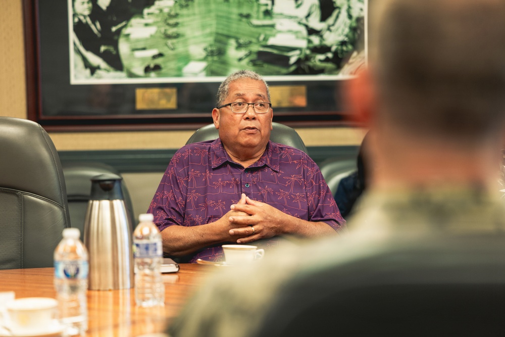 A middle-aged man wearing glasses and a purple Hawaiian-style shirt is seated at a conference table, appearing to speak or present to others in the room. The setting appears to be a formal meeting room with leather chairs, a wooden table, and a framed photograph or artwork on the wall behind him. Water bottles and a thermos are visible on the table, suggesting an organized meeting or briefing.