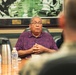 A middle-aged man wearing glasses and a purple Hawaiian-style shirt is seated at a conference table, appearing to speak or present to others in the room. The setting appears to be a formal meeting room with leather chairs, a wooden table, and a framed photograph or artwork on the wall behind him. Water bottles and a thermos are visible on the table, suggesting an organized meeting or briefing.