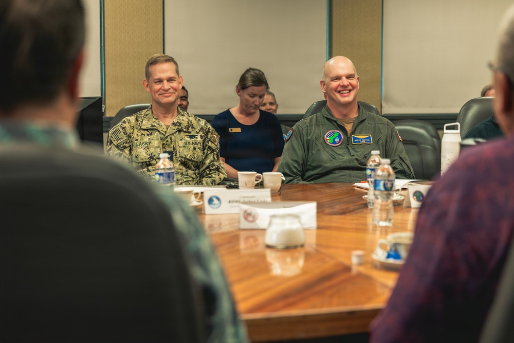 Two military personnel are seated at a conference table during what appears to be a formal meeting or briefing. The individual on the left wears U.S. Navy digital camouflage (NWU) with a name tape reading 'LASKY', while the individual on the right wears an olive green flight suit with patches. Both are smiling, suggesting a positive or lighthearted moment during the proceedings.