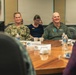 Two military personnel are seated at a conference table during what appears to be a formal meeting or briefing. The individual on the left wears U.S. Navy digital camouflage (NWU) with a name tape reading 'LASKY', while the individual on the right wears an olive green flight suit with patches. Both are smiling, suggesting a positive or lighthearted moment during the proceedings.