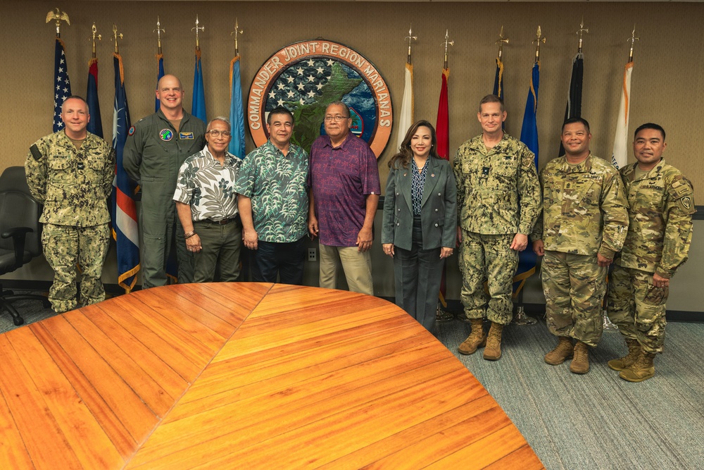 A group of nine individuals pose for a formal photograph in front of the 'Commander Joint Region Marianas' seal/emblem. The group includes both military personnel in various uniforms (Navy NWU, Army OCP, and flight suit) and civilians in casual attire. The setting appears to be an official conference or briefing room with multiple military and regional flags displayed.