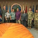 A group of nine individuals pose for a formal photograph in front of the 'Commander Joint Region Marianas' seal/emblem. The group includes both military personnel in various uniforms (Navy NWU, Army OCP, and flight suit) and civilians in casual attire. The setting appears to be an official conference or briefing room with multiple military and regional flags displayed.