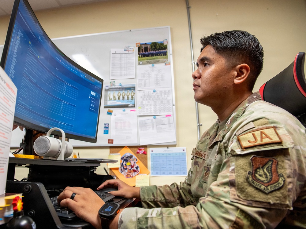 A U.S. Air Force service member wearing OCP (Operational Camouflage Pattern) uniform with a 'JA' (Judge Advocate) patch is working at a desk with a large curved monitor. The individual appears to be reviewing or inputting data into a software application displayed on the screen. The setting is an office environment with reference materials and documents posted on a bulletin board behind them.