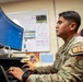 A U.S. Air Force service member wearing OCP (Operational Camouflage Pattern) uniform with a 'JA' (Judge Advocate) patch is working at a desk with a large curved monitor. The individual appears to be reviewing or inputting data into a software application displayed on the screen. The setting is an office environment with reference materials and documents posted on a bulletin board behind them.