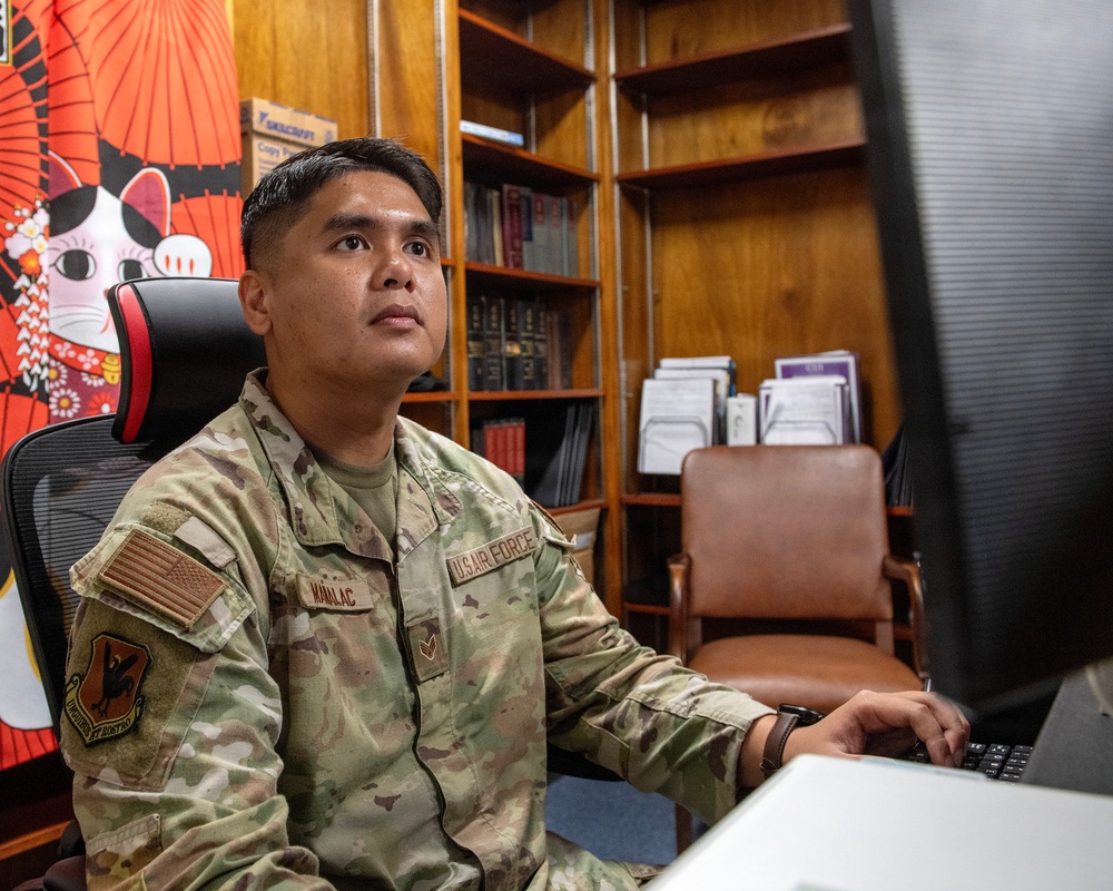 A U.S. Air Force airman in OCP (Operational Camouflage Pattern) uniform is seated at a desk working at a computer workstation. The name tape reads 'MANLAC' and the branch tape reads 'U.S. AIR FORCE,' with an American flag patch and a unit patch visible on the left sleeve. The setting appears to be an office environment with wooden bookshelves, reference materials, and Japanese-themed decorative items in the background.