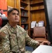 A U.S. Air Force airman in OCP (Operational Camouflage Pattern) uniform is seated at a desk working at a computer workstation. The name tape reads 'MANLAC' and the branch tape reads 'U.S. AIR FORCE,' with an American flag patch and a unit patch visible on the left sleeve. The setting appears to be an office environment with wooden bookshelves, reference materials, and Japanese-themed decorative items in the background.