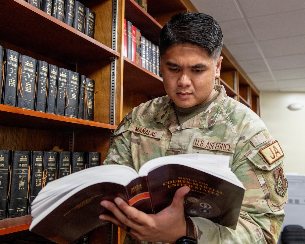 A U.S. Air Force service member named Mañalac, wearing OCP (Operational Camouflage Pattern) uniform with a 'JA' (Judge Advocate) patch, is reading the 2024 Manual for Courts-Martial United States in what appears to be a military legal library. The shelves behind him contain numerous bound legal volumes, many appearing to be Japanese-language legal texts, suggesting the photo was taken at a base in Japan. The image conveys a professional legal/JAG office setting.