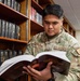 A U.S. Air Force service member named Mañalac, wearing OCP (Operational Camouflage Pattern) uniform with a 'JA' (Judge Advocate) patch, is reading the 2024 Manual for Courts-Martial United States in what appears to be a military legal library. The shelves behind him contain numerous bound legal volumes, many appearing to be Japanese-language legal texts, suggesting the photo was taken at a base in Japan. The image conveys a professional legal/JAG office setting.