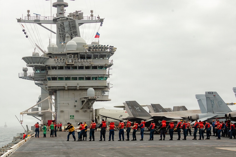 The image shows the flight deck of a U.S. Navy aircraft carrier with a large group of sailors conducting what appears to be a foreign object debris (FOD) walkdown or safety sweep. Multiple F/A-18 Hornet/Super Hornet aircraft are parked on the starboard side of the deck, and the carrier's island superstructure is prominently visible on the left. The scene takes place under overcast skies at sea.