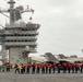 The image shows the flight deck of a U.S. Navy aircraft carrier with a large group of sailors conducting what appears to be a foreign object debris (FOD) walkdown or safety sweep. Multiple F/A-18 Hornet/Super Hornet aircraft are parked on the starboard side of the deck, and the carrier's island superstructure is prominently visible on the left. The scene takes place under overcast skies at sea.