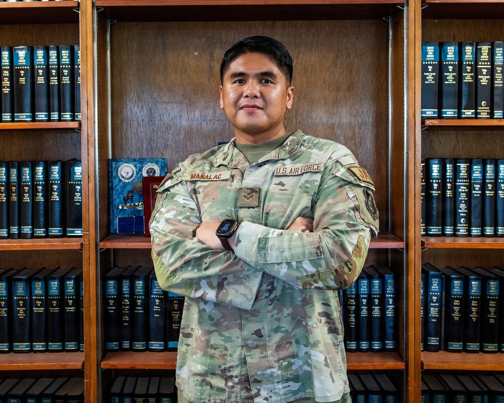 A U.S. Air Force service member named Mañalac stands with arms crossed in front of a wooden bookshelf filled with legal volumes (USCS - United States Code Service). The individual is wearing the standard OCP (Operational Camouflage Pattern) uniform with visible rank insignia and a smartwatch. The setting appears to be a military legal office or law library.