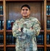 A U.S. Air Force service member named Mañalac stands with arms crossed in front of a wooden bookshelf filled with legal volumes (USCS - United States Code Service). The individual is wearing the standard OCP (Operational Camouflage Pattern) uniform with visible rank insignia and a smartwatch. The setting appears to be a military legal office or law library.