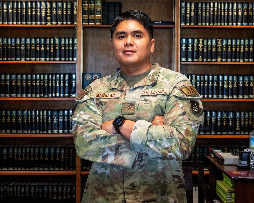 A U.S. Air Force service member named Mañalac stands with arms crossed in front of a large wooden bookshelf filled with uniform dark-bound volumes, likely a law library. The individual wears an Operational Camouflage Pattern (OCP) uniform with visible name tape, branch tape, rank insignia, and a 'JAG' (Judge Advocate General) patch. The setting and patch suggest this is a military legal office environment.