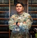 A U.S. Air Force service member named Mañalac stands with arms crossed in front of a large wooden bookshelf filled with uniform dark-bound volumes, likely a law library. The individual wears an Operational Camouflage Pattern (OCP) uniform with visible name tape, branch tape, rank insignia, and a 'JAG' (Judge Advocate General) patch. The setting and patch suggest this is a military legal office environment.