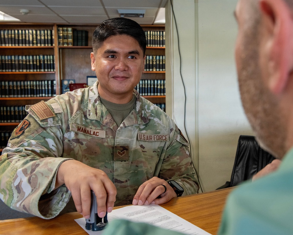 A U.S. Air Force servicemember named Mañalac, wearing OCP (Operational Camouflage Pattern) uniform, is seated at a wooden desk across from a civilian or another military member. He is holding a stamp/seal over a document, suggesting a notarial or administrative function. The setting appears to be a legal office or library, with rows of bound volumes visible in the background.
