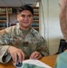 A U.S. Air Force servicemember named Mañalac, wearing OCP (Operational Camouflage Pattern) uniform, is seated at a wooden desk across from a civilian or another military member. He is holding a stamp/seal over a document, suggesting a notarial or administrative function. The setting appears to be a legal office or library, with rows of bound volumes visible in the background.