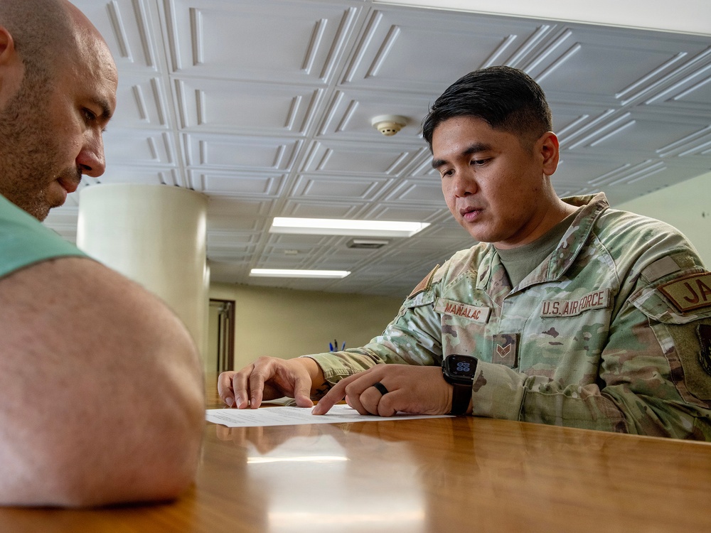 A U.S. Air Force servicemember in OCP (Operational Camouflage Pattern) uniform is seated at a wooden table reviewing a document with a civilian. The servicemember, whose name tape reads 'MAÑALAC,' is pointing to specific content on the paper while appearing to explain or discuss it. The setting appears to be an indoor administrative or office environment.