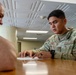 A U.S. Air Force servicemember in OCP (Operational Camouflage Pattern) uniform is seated at a wooden table reviewing a document with a civilian. The servicemember, whose name tape reads 'MAÑALAC,' is pointing to specific content on the paper while appearing to explain or discuss it. The setting appears to be an indoor administrative or office environment.