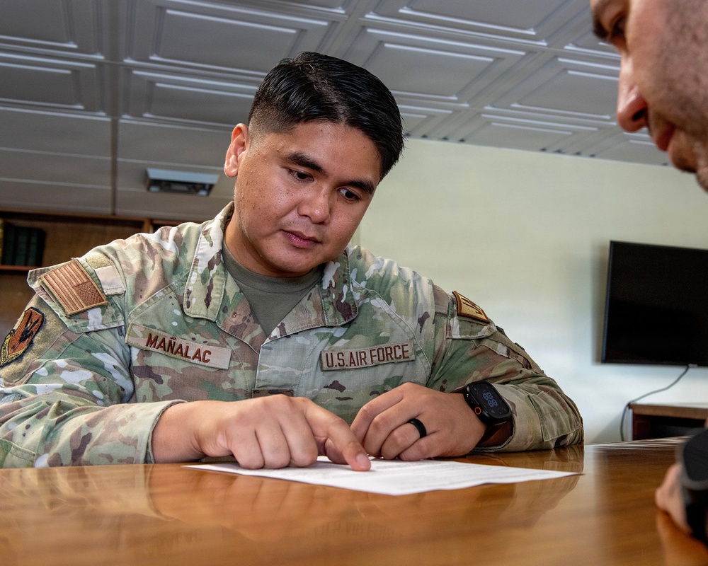 A U.S. Air Force service member named Mañalac is seated at a wooden table, pointing to a document while apparently reviewing or discussing its contents with another person. The airman is wearing an Operational Camouflage Pattern (OCP) uniform with a U.S. flag patch and unit insignia. The setting appears to be an indoor office or administrative space.