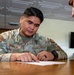 A U.S. Air Force service member named Mañalac is seated at a wooden table, pointing to a document while apparently reviewing or discussing its contents with another person. The airman is wearing an Operational Camouflage Pattern (OCP) uniform with a U.S. flag patch and unit insignia. The setting appears to be an indoor office or administrative space.