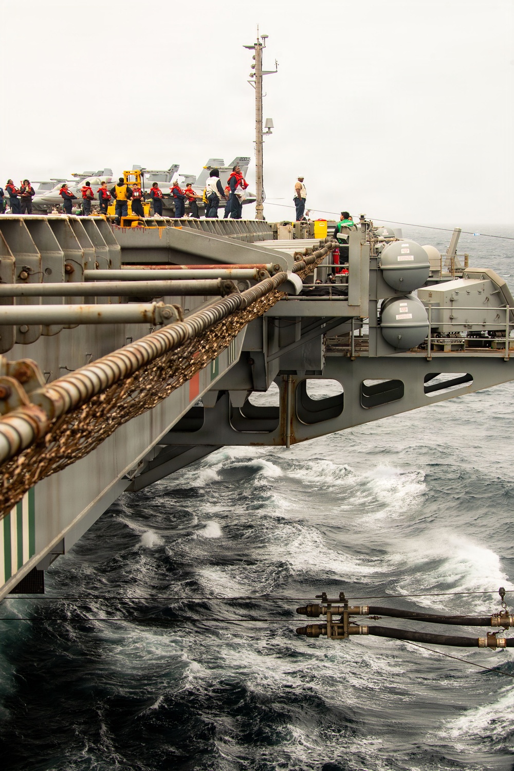 The image shows a replenishment-at-sea (RAS) operation between two naval vessels underway in rough ocean conditions. Transfer lines, fuel hoses, and tensioned cables are strung between the ships, with numerous crew members visible on the flight deck wearing red and yellow safety vests. Aircraft, likely F/A-18 Hornets, are visible parked on the flight deck of what appears to be a US Navy aircraft carrier.
