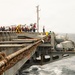 The image shows a replenishment-at-sea (RAS) operation between two naval vessels underway in rough ocean conditions. Transfer lines, fuel hoses, and tensioned cables are strung between the ships, with numerous crew members visible on the flight deck wearing red and yellow safety vests. Aircraft, likely F/A-18 Hornets, are visible parked on the flight deck of what appears to be a US Navy aircraft carrier.