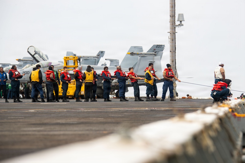 A group of naval personnel are conducting a line-handling or tug-of-war drill on the flight deck of a US Navy aircraft carrier. Multiple sailors wearing red life vests and blue coveralls are pulling on a rope or line in a coordinated formation. Several F/A-18 Hornet or Super Hornet jet aircraft are visible in the background, with modex numbers '200' visible on at least two aircraft.