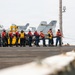 A group of naval personnel are conducting a line-handling or tug-of-war drill on the flight deck of a US Navy aircraft carrier. Multiple sailors wearing red life vests and blue coveralls are pulling on a rope or line in a coordinated formation. Several F/A-18 Hornet or Super Hornet jet aircraft are visible in the background, with modex numbers '200' visible on at least two aircraft.
