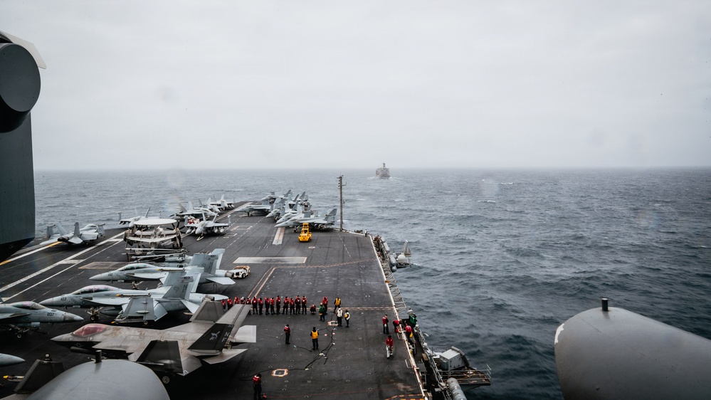 An aerial view from the island superstructure of a large aircraft carrier underway at sea in overcast conditions. The flight deck is populated with multiple fixed-wing jet aircraft, deck crew in colored jerseys, and support vehicles. A second naval vessel is visible on the horizon in the background.