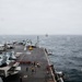 An aerial view from the island superstructure of a large aircraft carrier underway at sea in overcast conditions. The flight deck is populated with multiple fixed-wing jet aircraft, deck crew in colored jerseys, and support vehicles. A second naval vessel is visible on the horizon in the background.