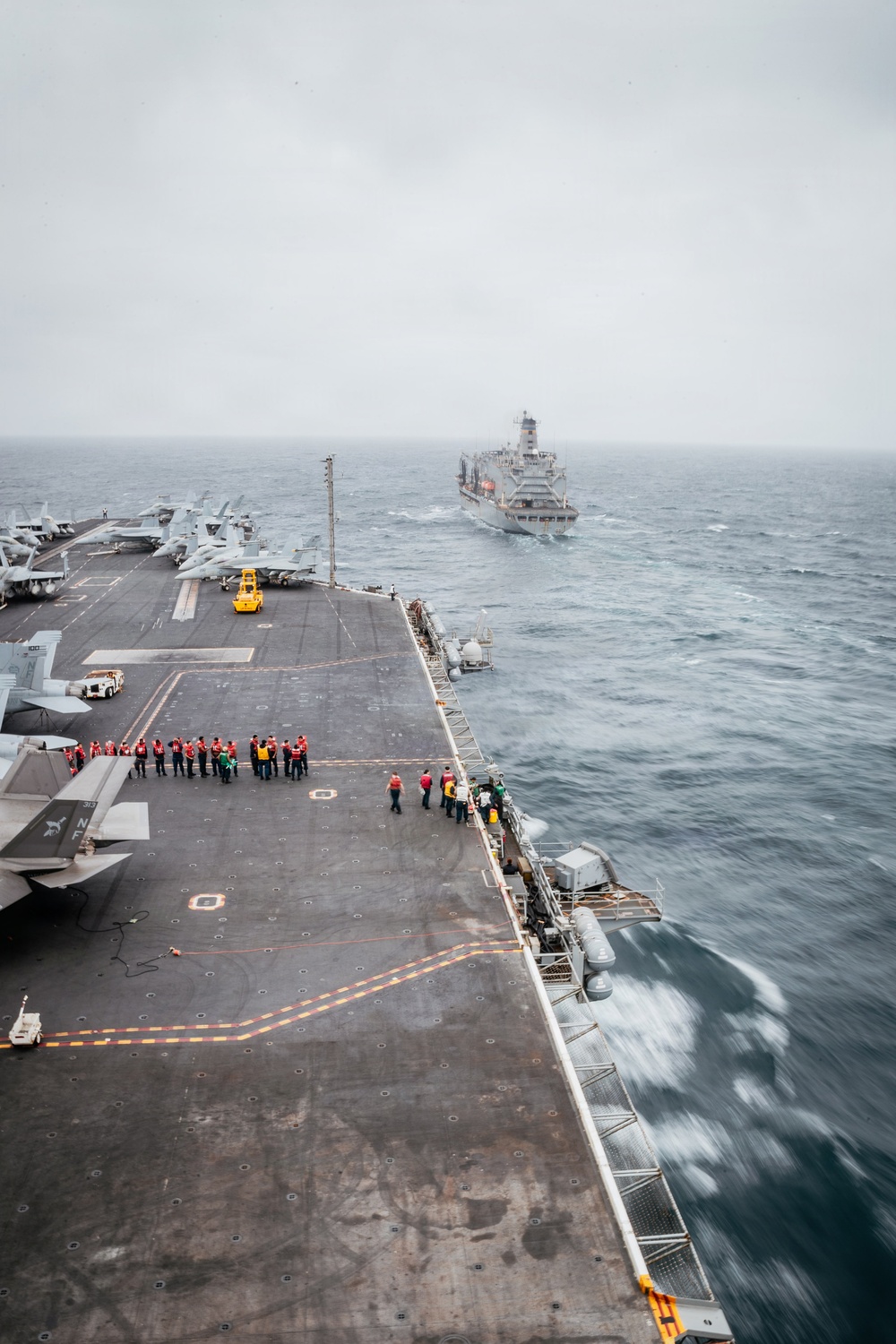 The image shows the flight deck of a large aircraft carrier underway at sea, with personnel gathered along the starboard edge, likely preparing for or conducting an underway replenishment (UNREP) operation. A replenishment vessel is visible in the background at a parallel course. Multiple fixed-wing aircraft are parked on the port side of the flight deck.