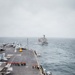 The image shows the flight deck of a large aircraft carrier underway at sea, with personnel gathered along the starboard edge, likely preparing for or conducting an underway replenishment (UNREP) operation. A replenishment vessel is visible in the background at a parallel course. Multiple fixed-wing aircraft are parked on the port side of the flight deck.